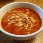 Close-up of a white bowl filled with vibrant red broth and shredded chicken for chicken pozole.