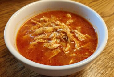 Close-up of a white bowl filled with vibrant red broth and shredded chicken for chicken pozole.