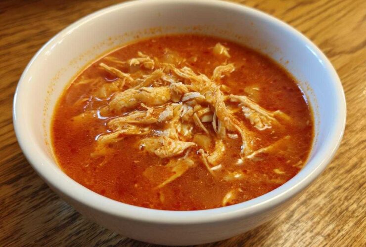 Close-up of a white bowl filled with vibrant red broth and shredded chicken for chicken pozole.