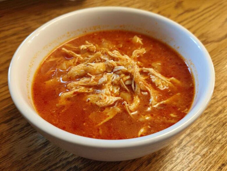 Close-up of a white bowl filled with vibrant red broth and shredded chicken for chicken pozole.