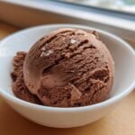A close-up of a scoop of rich chocolate protein ice cream topped with flaky sea salt in a white bowl.