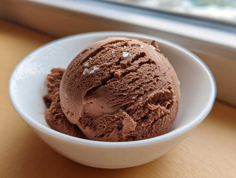 A close-up of a scoop of rich chocolate protein ice cream topped with flaky sea salt in a white bowl.
