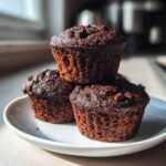 Three rich, dark chocolate zucchini muffins stacked on a white plate, highlighted by natural light.