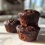 A stack of two moist chocolate zucchini muffins topped with chocolate chips, with a third muffin blurred in the background.