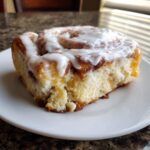 A single, fluffy serving of cinnamon roll casserole topped with thick white icing, resting on a white plate.