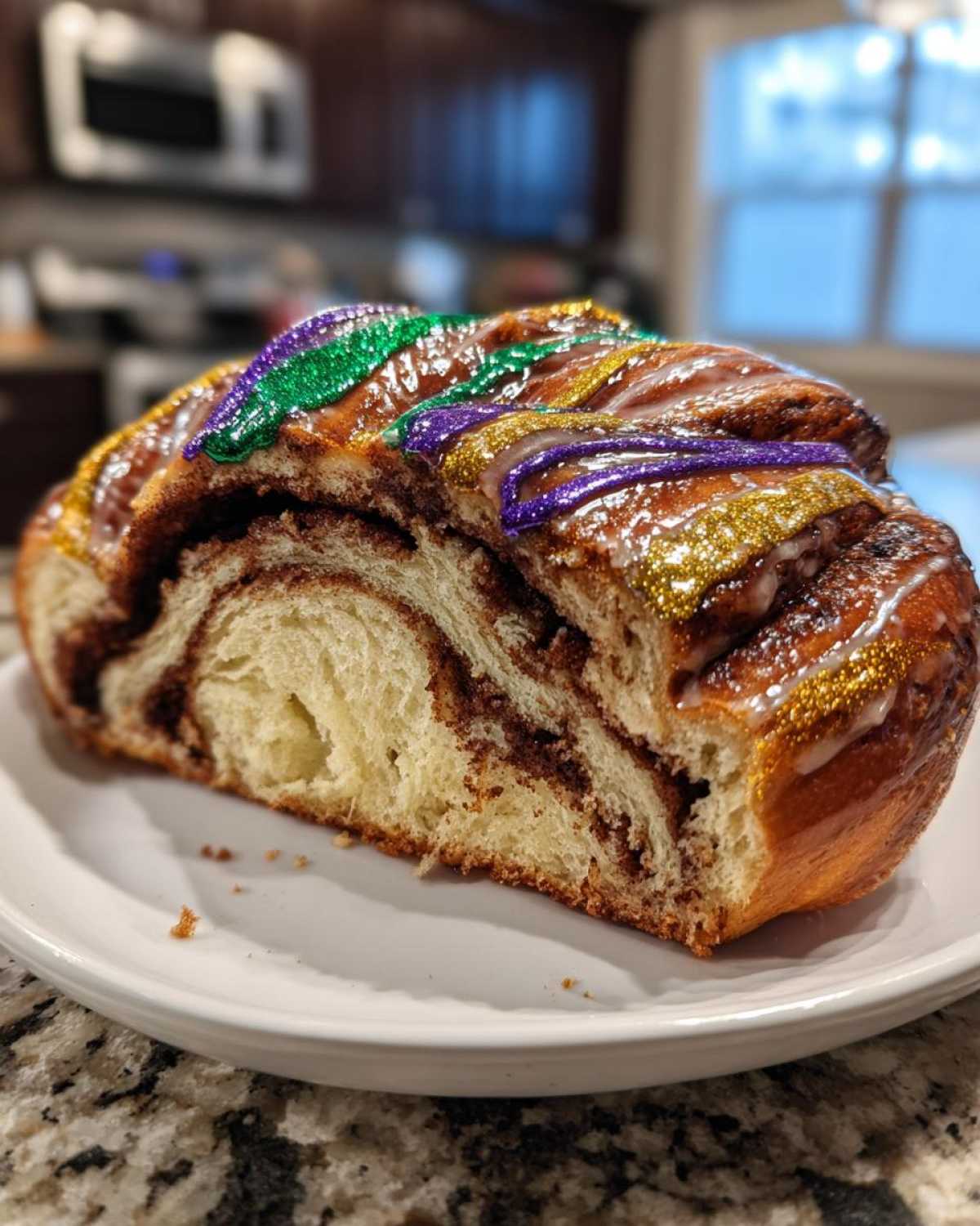 Cross-section of a rich, cinnamon-swirled king cake topped with purple, green, and gold sparkling icing.