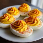 Close-up of five beautifully piped classic deviled eggs sprinkled with bright red paprika on a white plate.