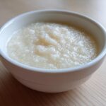 A close-up of creamy, pale coconut chia pudding served in a small white bowl on a light wooden surface.