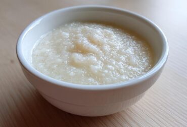A close-up of creamy, pale coconut chia pudding served in a small white bowl on a light wooden surface.