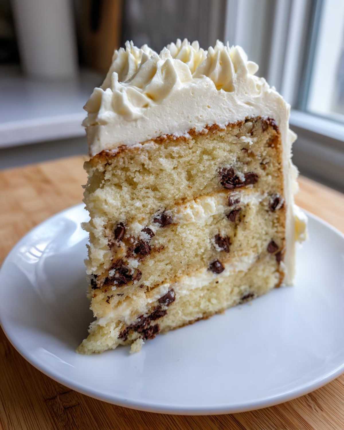 A thick slice of chocolate chip cookie cake recipe layers with vanilla frosting on a white plate.