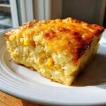 A golden-brown slice of moist cornbread casserole packed with whole corn kernels on a white plate.