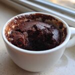 A rich, dark chocolate Cottage Cheese Microwave Brownie baked inside a white mug, sitting on a speckled countertop near a window.