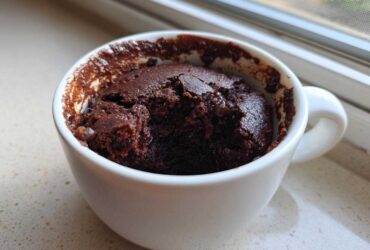 A rich, dark chocolate Cottage Cheese Microwave Brownie baked inside a white mug, sitting on a speckled countertop near a window.