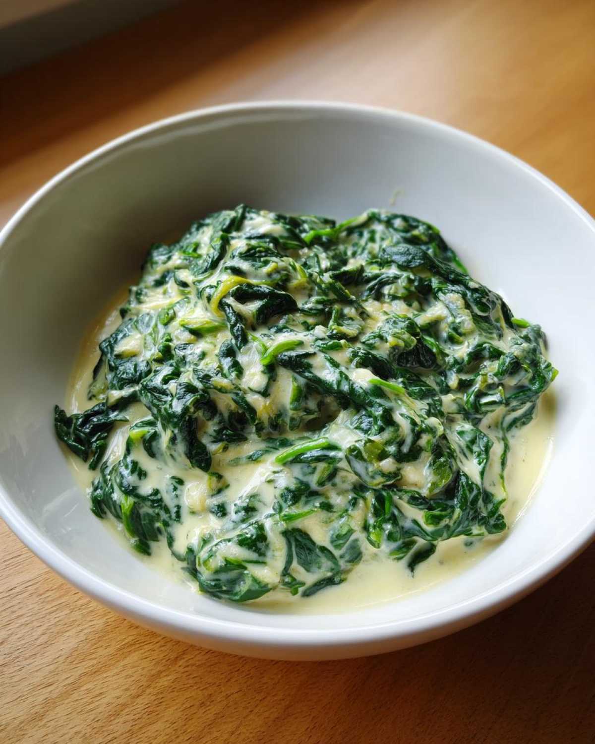 A close-up of rich, green creamed spinach recipe served in a white bowl on a wooden surface.