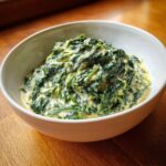 A close-up of a white bowl filled with rich, homemade creamed spinach recipe, served on a wooden surface.