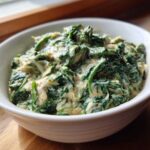 A white bowl filled with rich, green creamed spinach recipe, served on a wooden surface near a window.