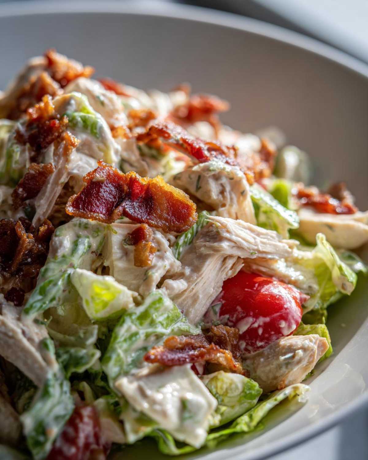 Close-up of a bowl of creamy blt chicken salad featuring shredded chicken, crisp lettuce, cherry tomatoes, and crispy bacon pieces.