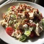 A bowl of creamy blt chicken salad featuring chunks of chicken, crisp lettuce, halved cherry tomatoes, and topped with crispy bacon.