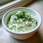A close-up of creamy, light green dill dip topped with fresh dill sprigs in a white serving bowl.