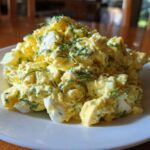A generous serving of creamy, yellow egg salad recipe garnished with fresh dill, presented on a white plate.