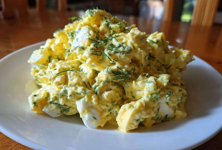 A generous serving of creamy, yellow egg salad recipe garnished with fresh dill, presented on a white plate.