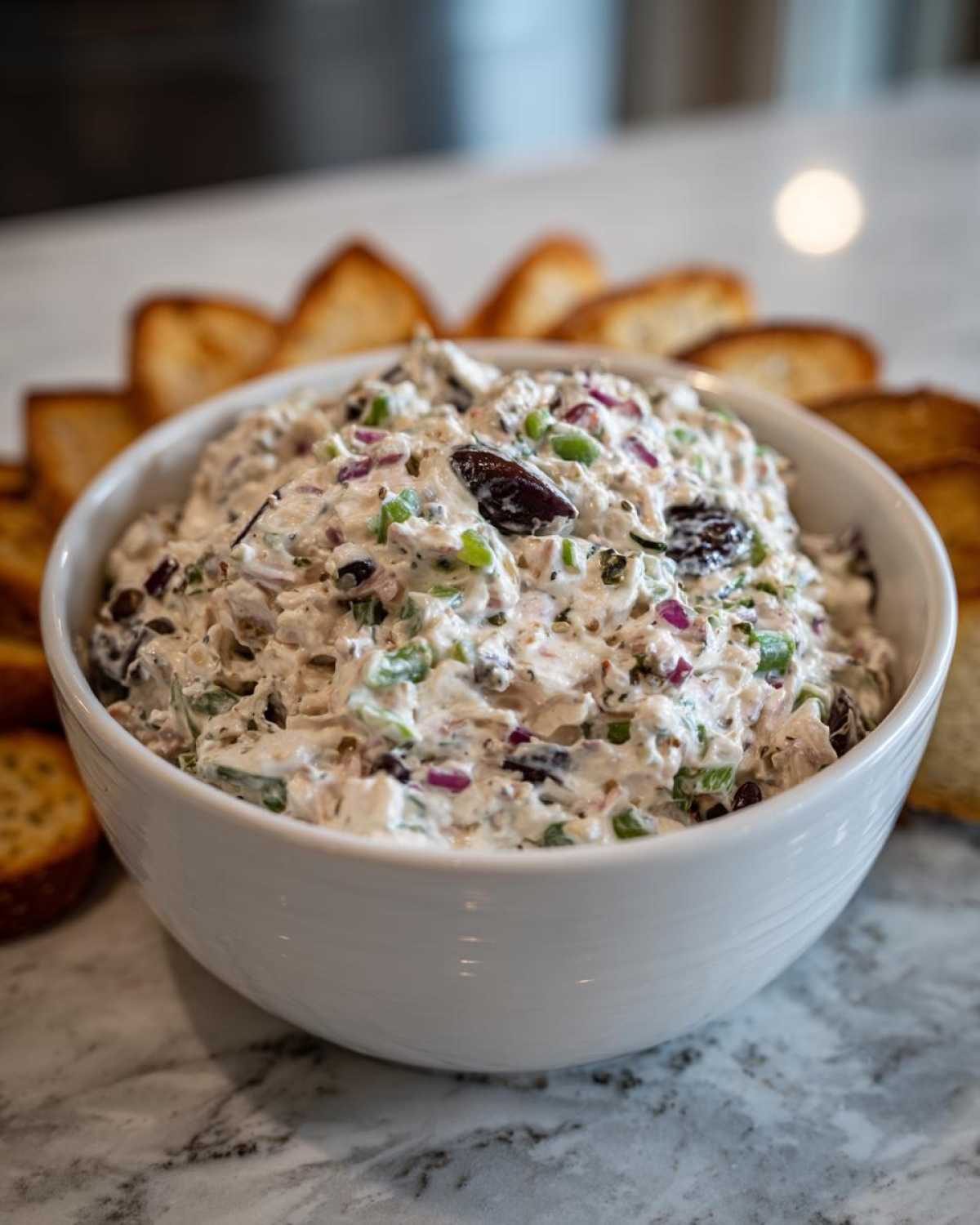 A white bowl filled with creamy hoagie dip, featuring visible chunks of meat, red onion, green peppers, and black olives, surrounded by toasted bread slices.