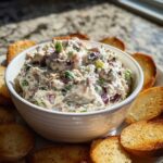A white bowl filled with creamy hoagie dip mixed with red onions and green onions, surrounded by toasted bread rounds.