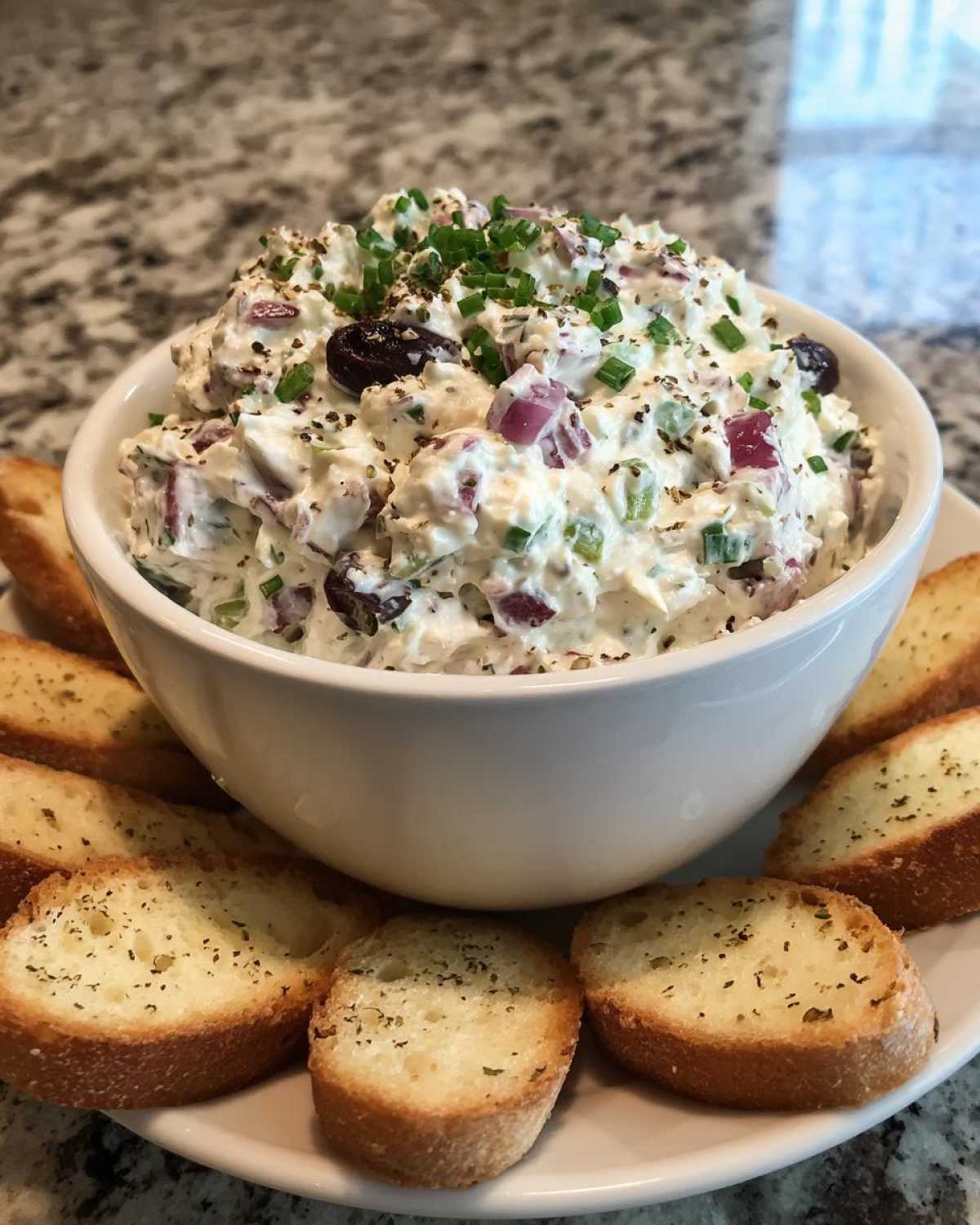 A white bowl filled with creamy hoagie dip, garnished with chives and olives, surrounded by toasted bread slices.