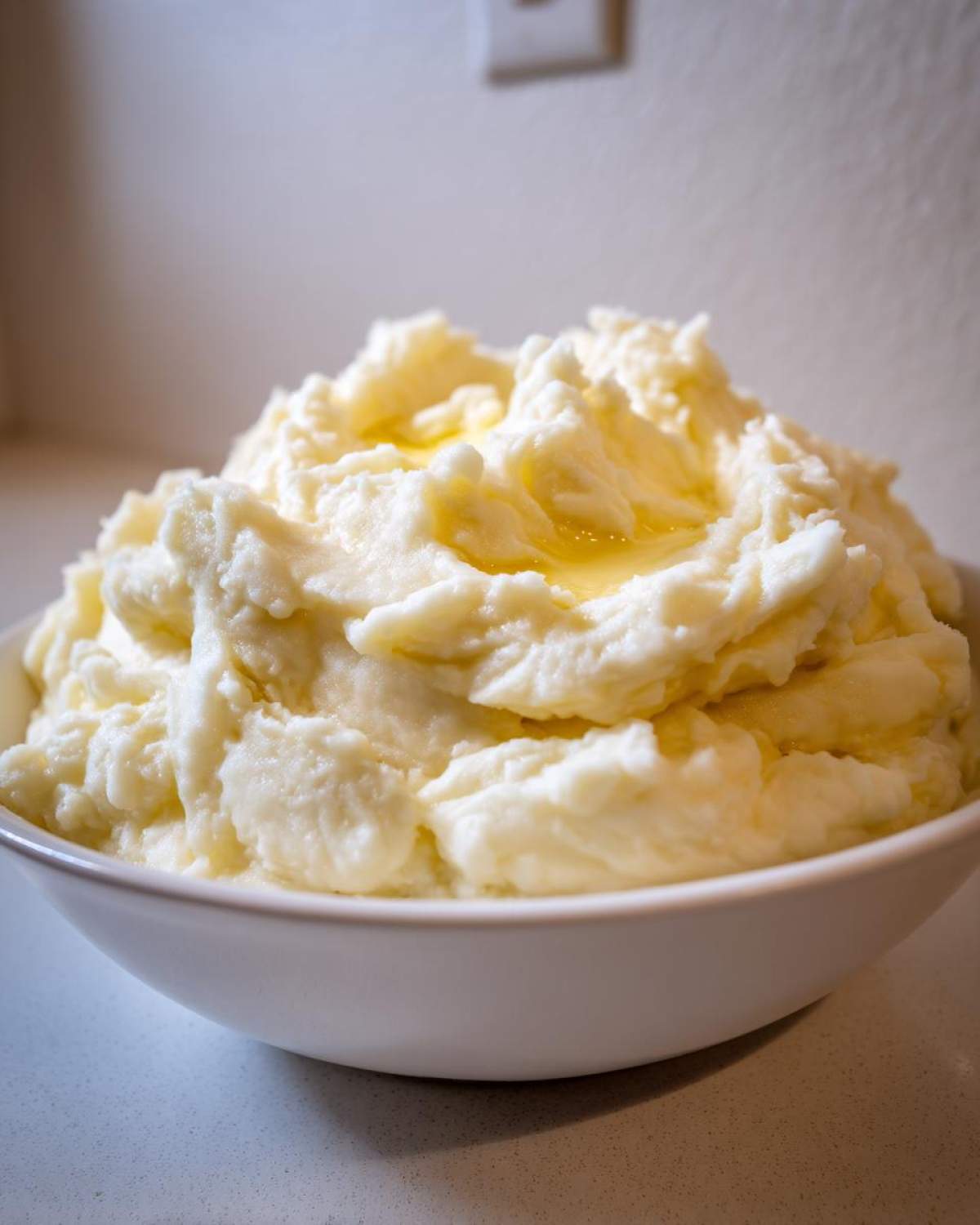 A large white bowl filled with fluffy, rich creamy mashed potatoes topped with melted butter.