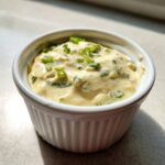 A close-up of creamy, yellow remoulade sauce topped with fresh chopped green onions in a white ramekin.
