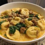 Close-up of a white bowl filled with creamy tortellini soup, featuring pasta, ground sausage, and wilted spinach.