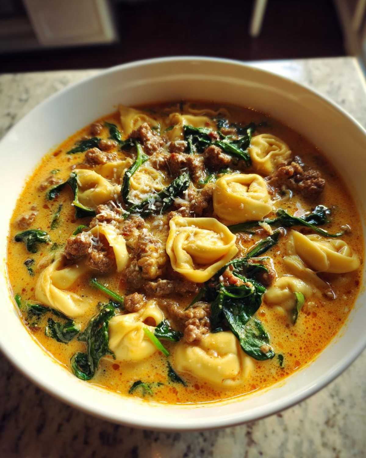 A close-up bowl of creamy tortellini soup featuring cheese tortellini, ground sausage, and wilted spinach.