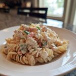 A close-up of a serving of creamy tuna pasta salad mixed with rotini pasta and chopped vegetables on a white plate.