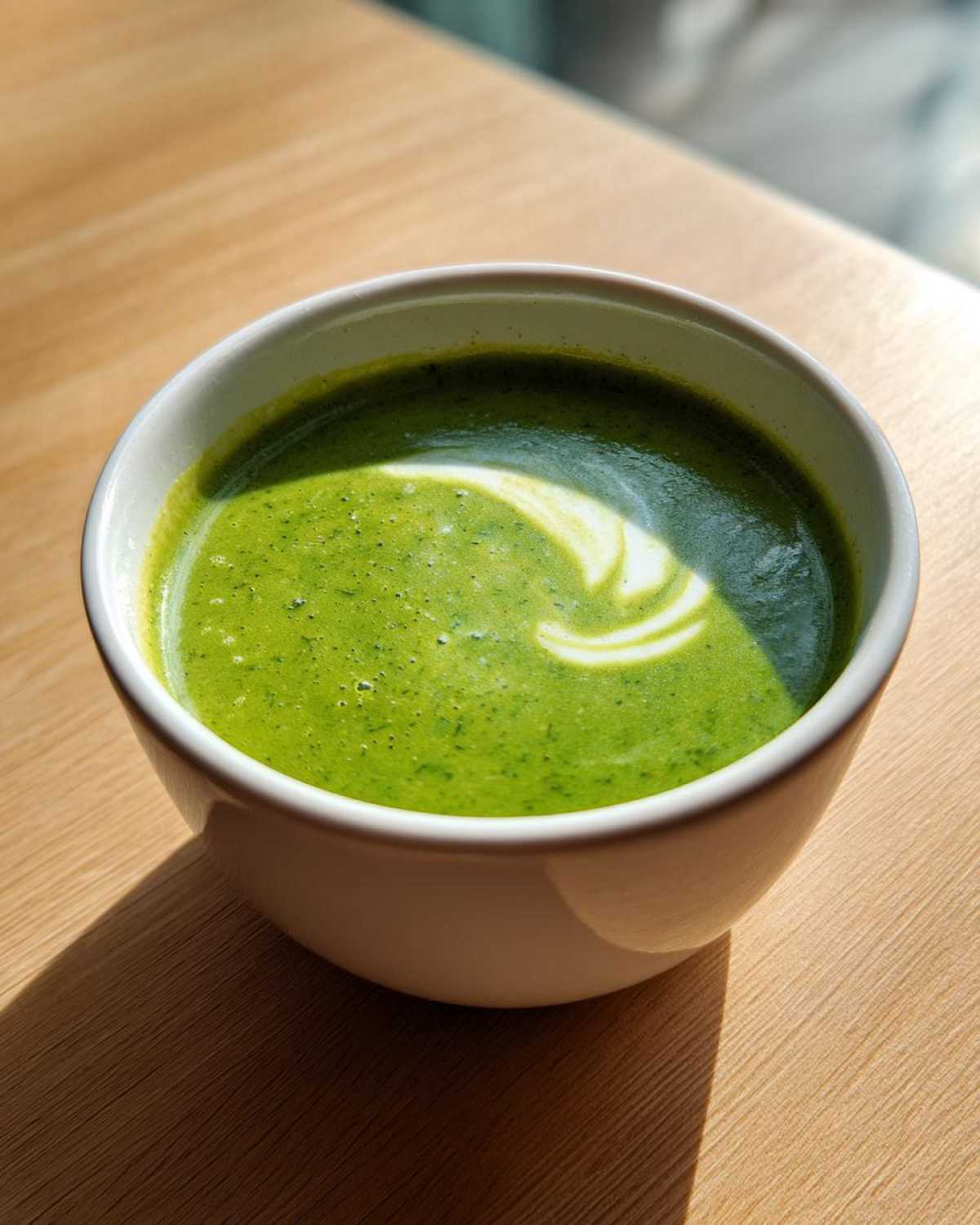 Close-up of a bowl of vibrant green zucchini soup, garnished with a white cream swirl, sitting on a wooden table.