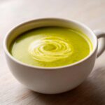 Close-up of vibrant green zucchini soup topped with a swirl of cream in a white mug.