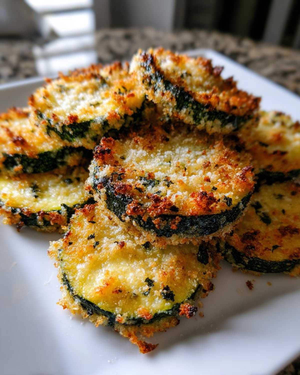Close-up of several golden brown, crispy baked parmesan zucchini slices stacked on a white plate.