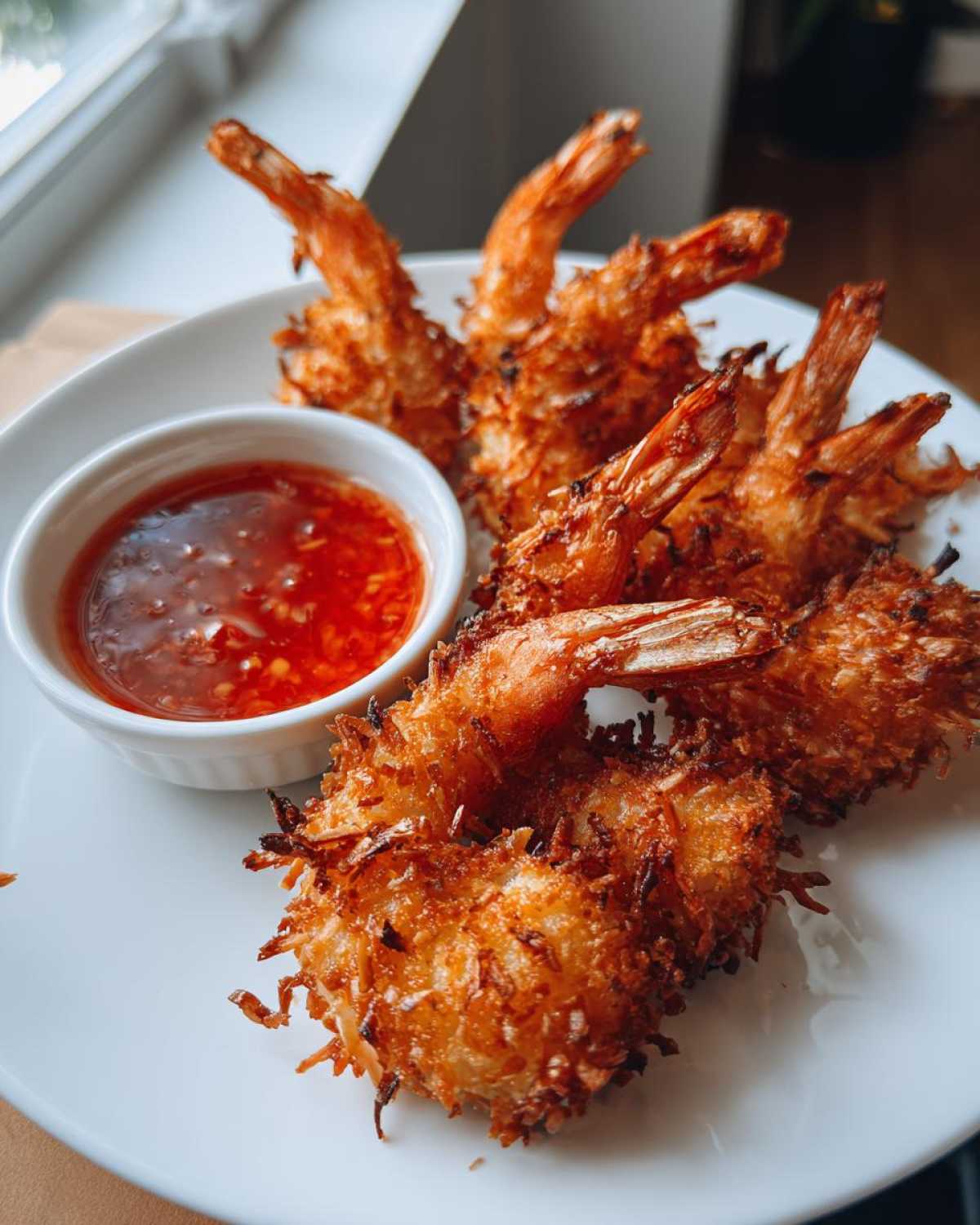 Close-up of golden brown, crispy coconut shrimp served on a white plate next to a small bowl of red sweet chili sauce.