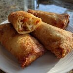 Four golden brown, crispy fried egg rolls on a white plate, one cut open showing cabbage filling.