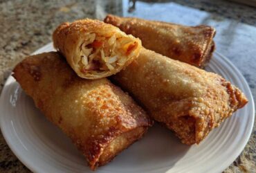 Four golden brown, crispy fried egg rolls on a white plate, one cut open showing cabbage filling.