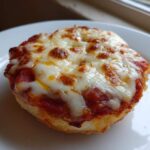 A close-up of a single, crispy english muffin pizza topped with bubbling tomato sauce and melted, slightly browned mozzarella cheese.
