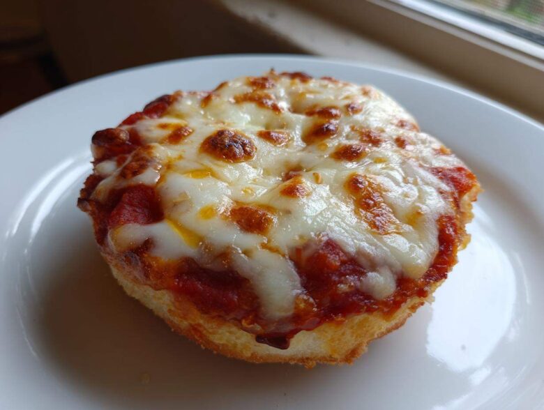 A close-up of a single, crispy english muffin pizza topped with bubbling tomato sauce and melted, slightly browned mozzarella cheese.
