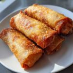 Close-up of four perfectly golden brown, crispy fried egg roll appetizers stacked on a white plate.