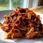 A close-up of a generous pile of saucy, shredded crock pot pulled pork served on a white plate.