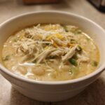 A close-up of a white bowl filled with creamy crockpot white chicken chili, topped with shredded chicken, cheese, and green onions.