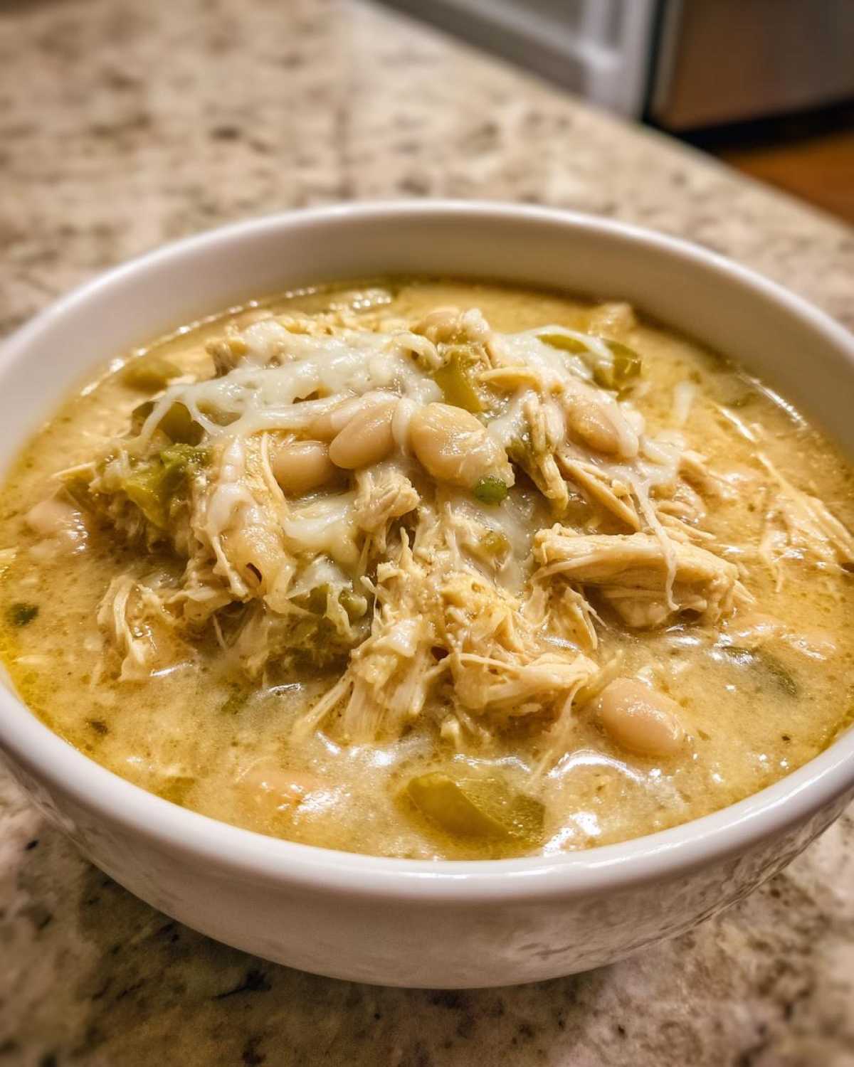 A close-up of a bowl of creamy crockpot white chicken chili topped with shredded chicken, white beans, and melted cheese.