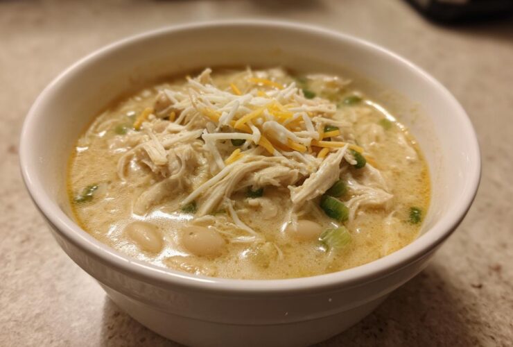 A close-up of a white bowl filled with creamy crockpot white chicken chili, topped with shredded chicken, cheese, and green onions.