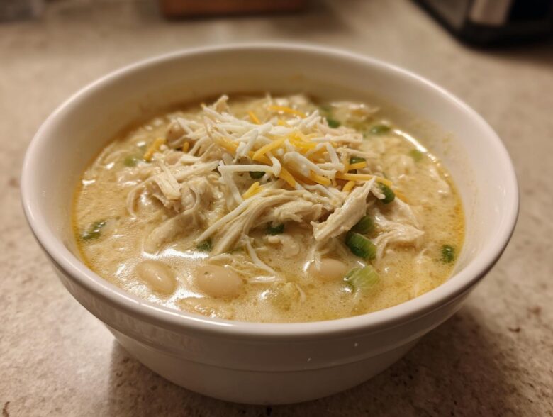 A close-up of a white bowl filled with creamy crockpot white chicken chili, topped with shredded chicken, cheese, and green onions.