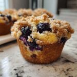 A close-up of a golden brown blueberry muffin featuring a generous, sugary crumb topping and visible baked blueberries.