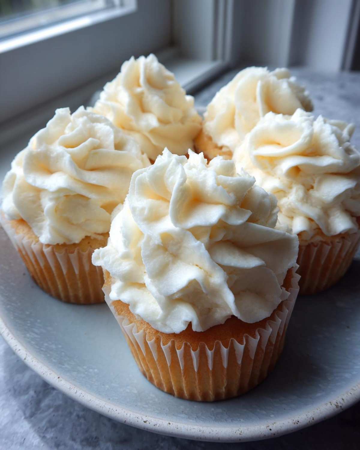 Four vanilla cupcakes topped high with swirls of rich vanilla buttercream frosting on a light blue plate.