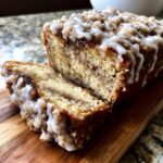 A close-up of a sliced coffee cake loaf showing the cinnamon swirl interior, crumb topping, and white icing drizzle.